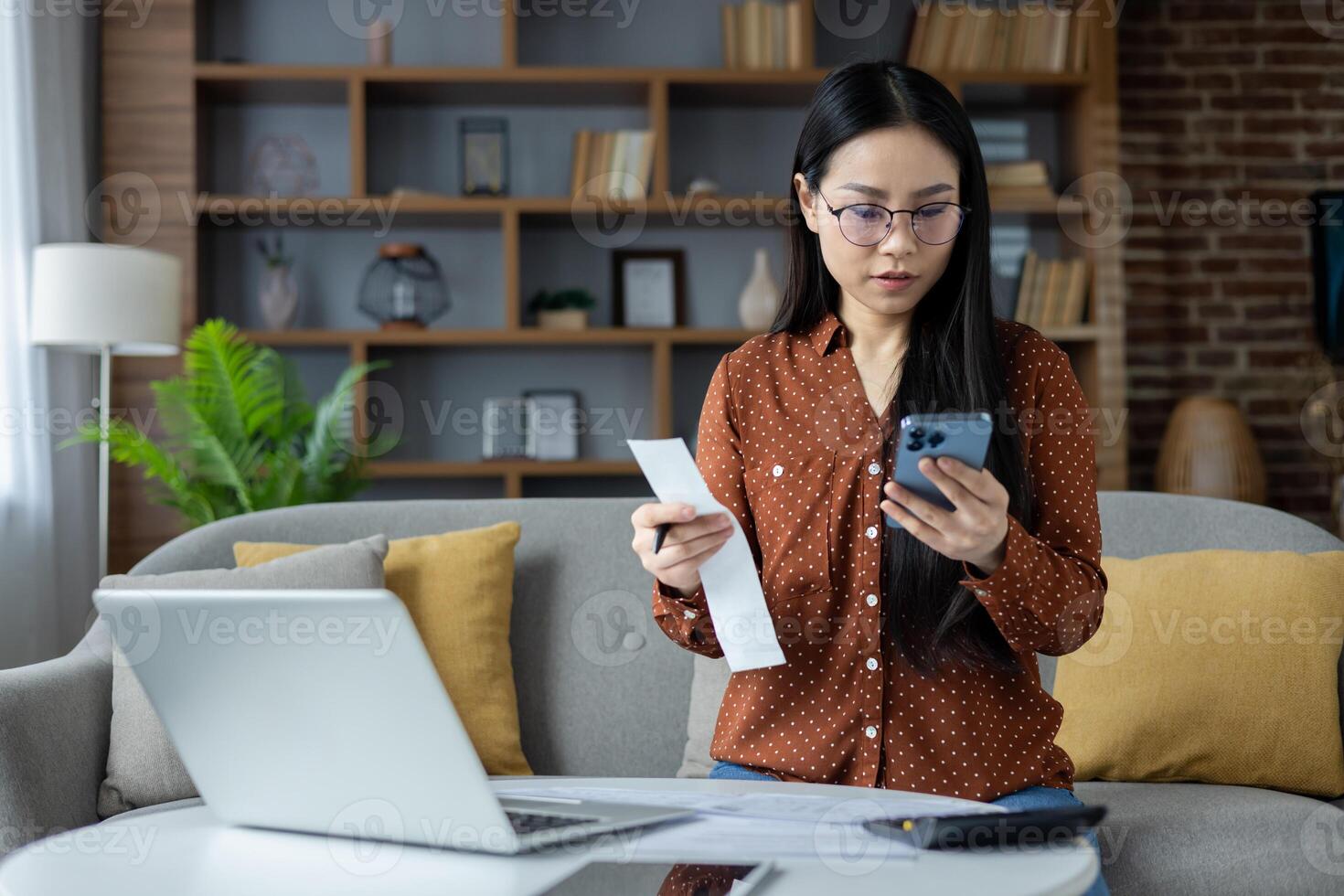 Young asian woman using phone for budget management. Sitting at home, she reviews bills with focused attention. Laptop and documents spread across table. photo