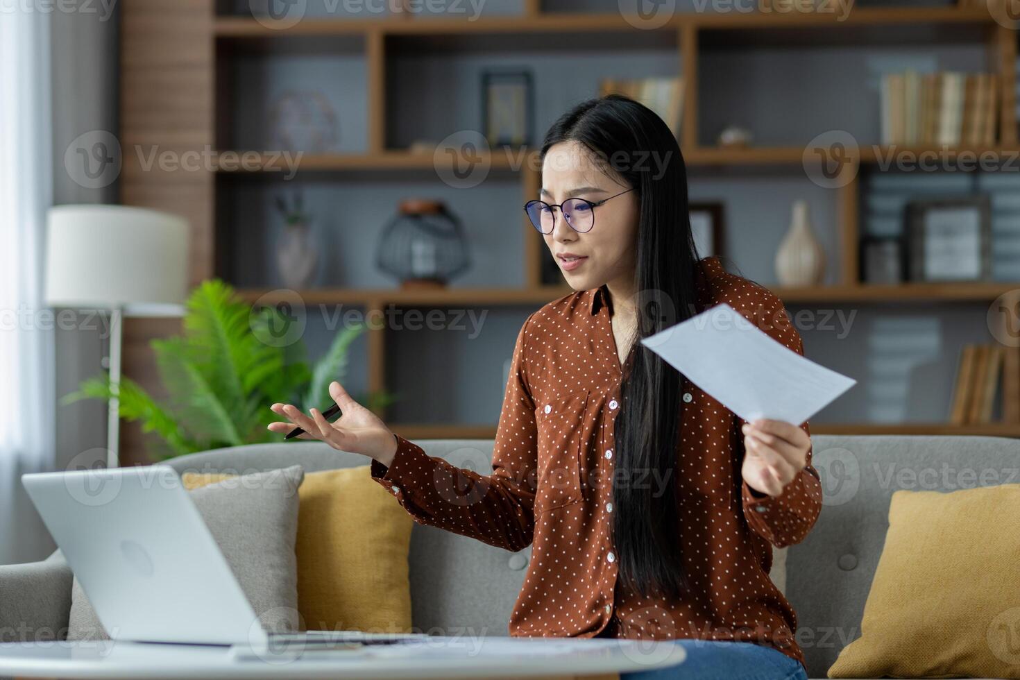 Asian woman in home office setting, engaged in call holding paper, discussing project. Laptop on table, remote work environment. Emblematic of modern communication and indoor workspace. photo