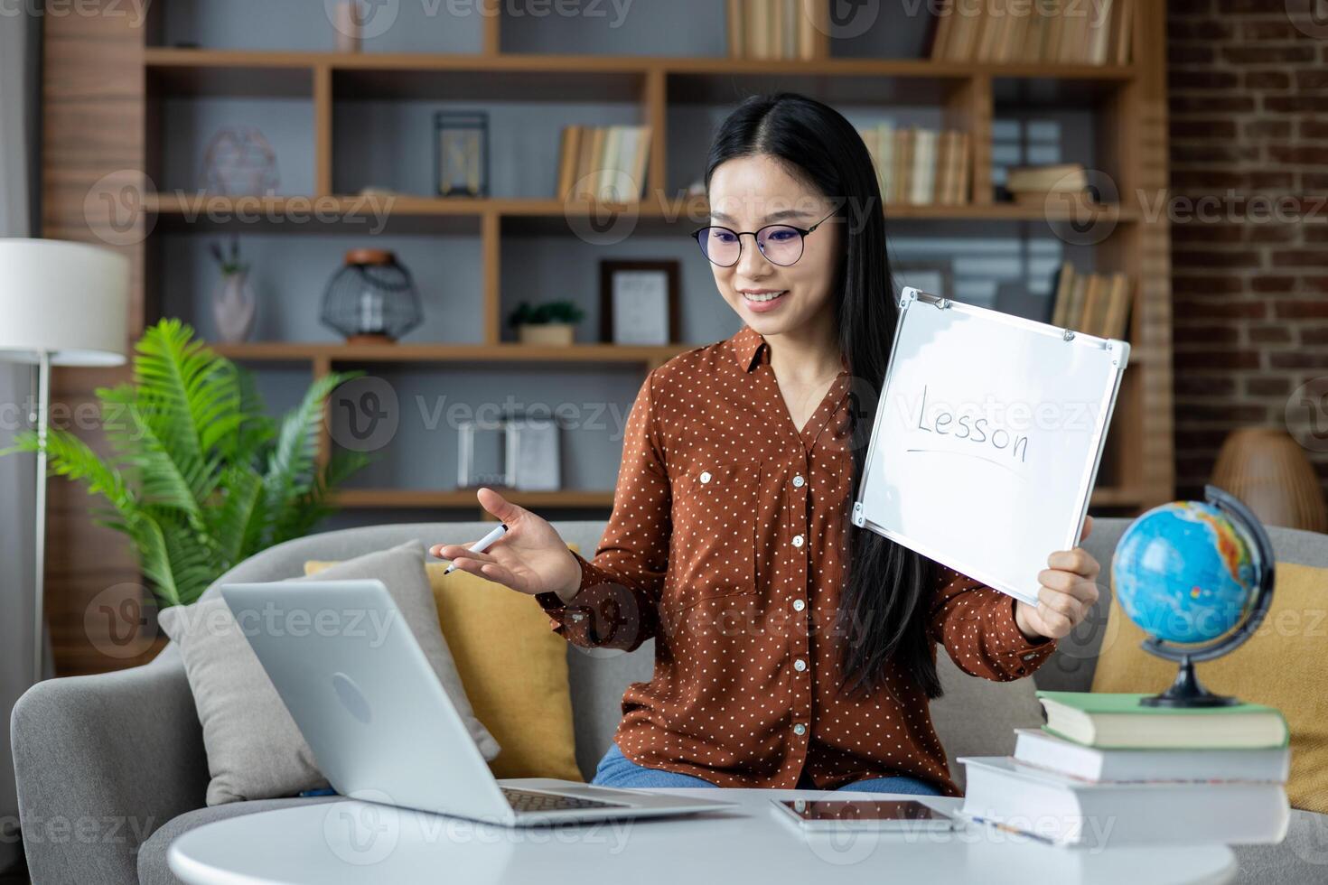 Asian woman conducting online lesson from home. She sits on sofa with laptop, holding a whiteboard marked Lesson, engaged in interactive teaching session, conveying education and technology. photo