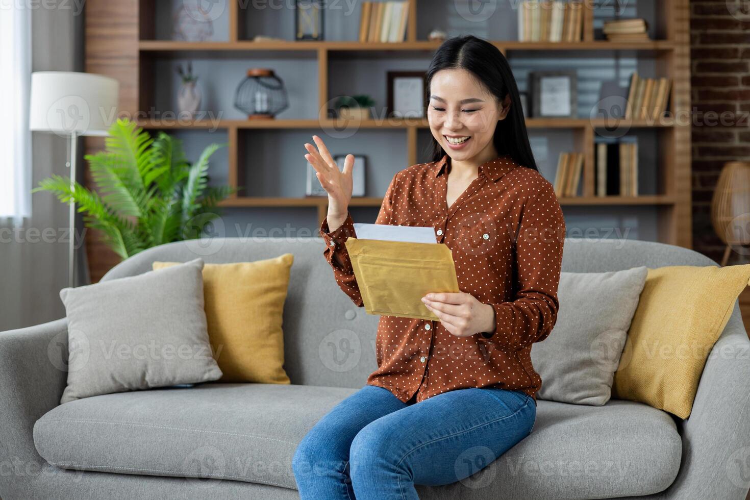 Asian woman sitting on cozy couch reading letter with happy expression. Comfortable home setting conveys joy and surprise as she receives message. Warm and inviting interior. photo