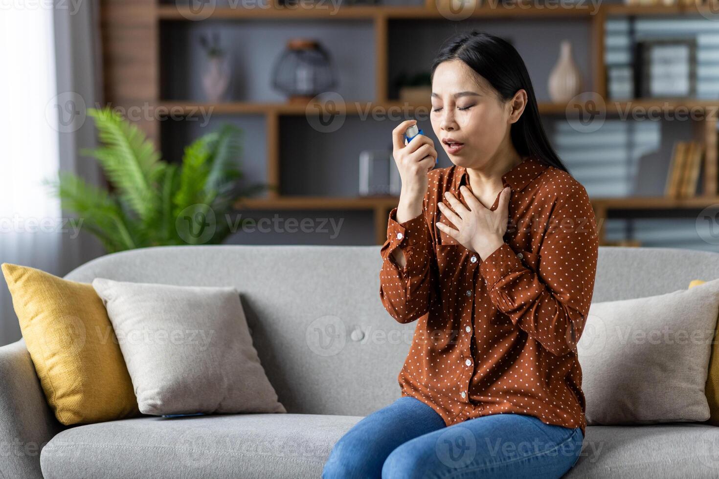 Asian woman using asthma inhaler, sitting on sofa experiencing breathing issue. Wearing polka dot blouse, managing respiratory health at home. Focused, calming atmosphere. photo