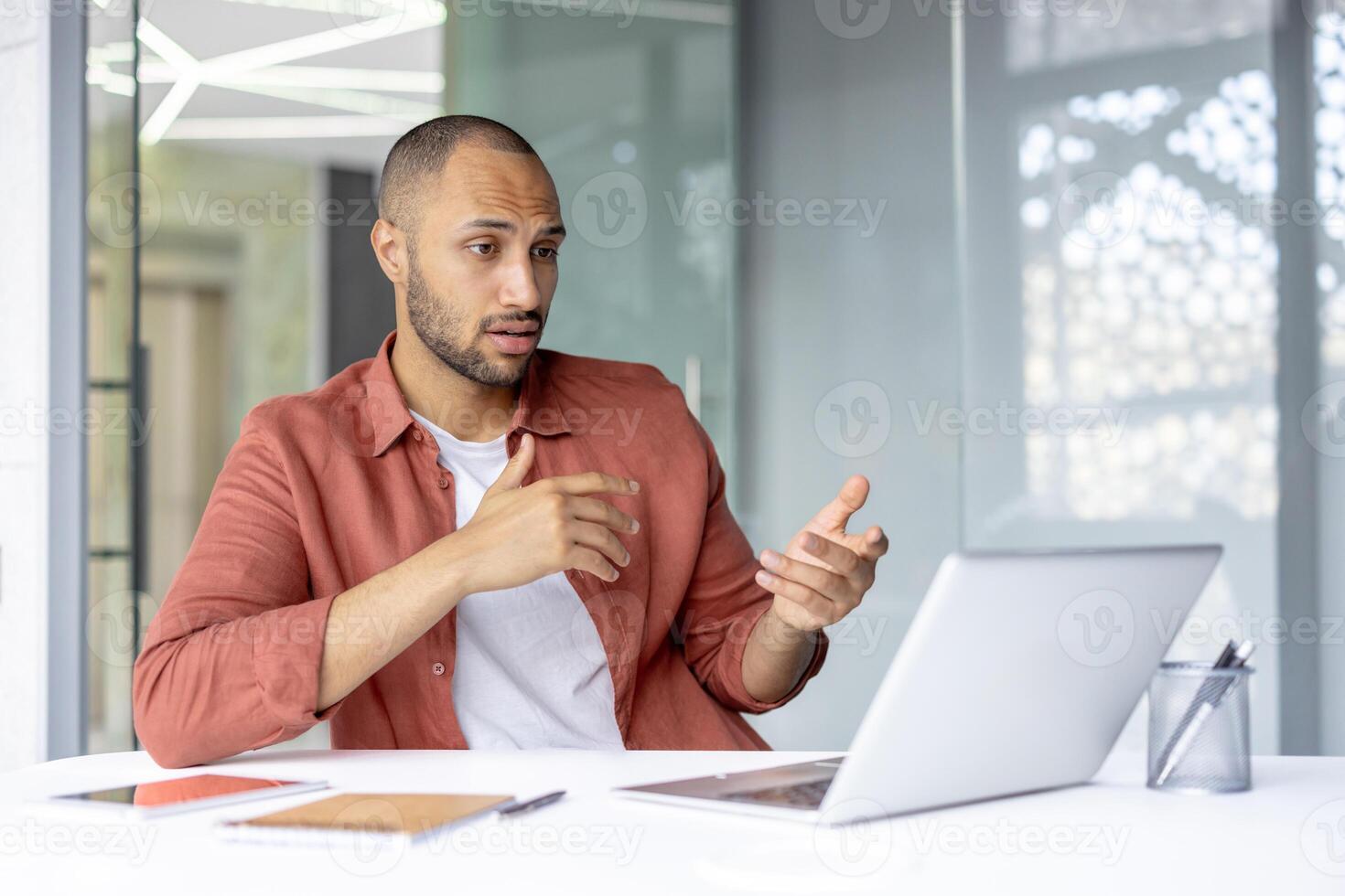 Serious man talking remotely with colleagues sitting at workplace inside office, office worker on online business meeting remote meeting with colleagues using laptop for call. photo