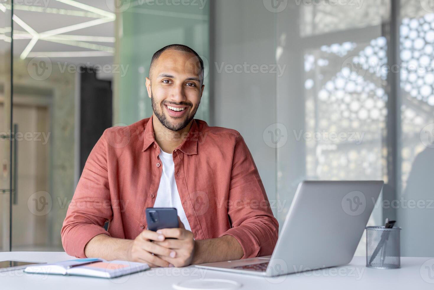 Portrait of a young businessman with a phone in his hands, a man works inside an office at a workplace with a laptop, looking joyfully at the camera. Office worker in casual clothes. photo