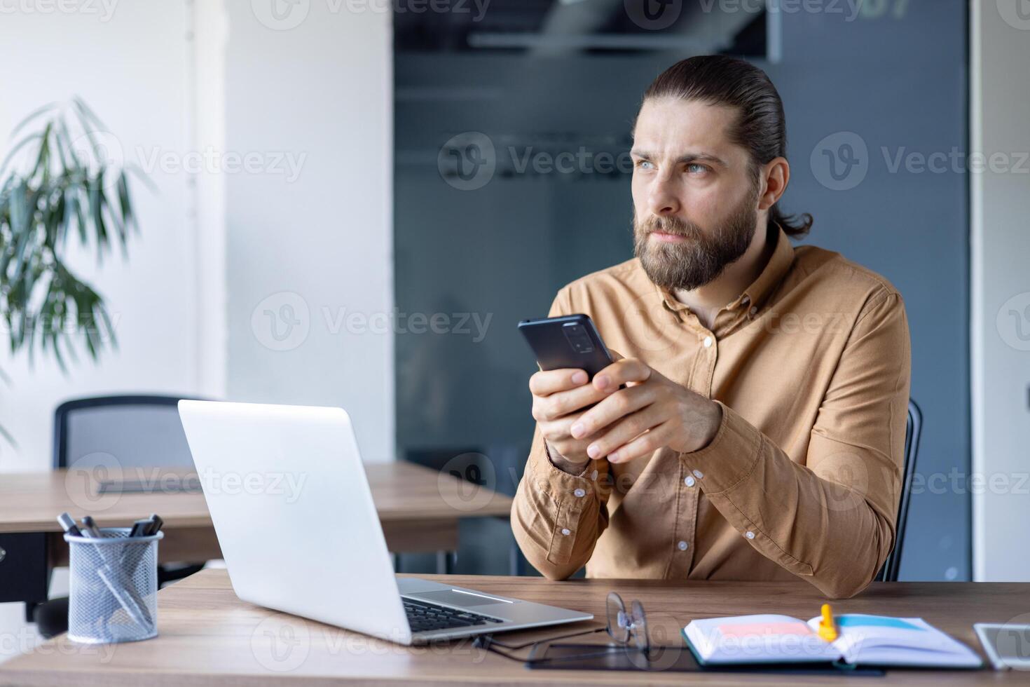 Focused man holds smartphone while working at an office desk with a laptop and documents. The office environment suggests analyzing, planning, and staying connected within a workplace. photo