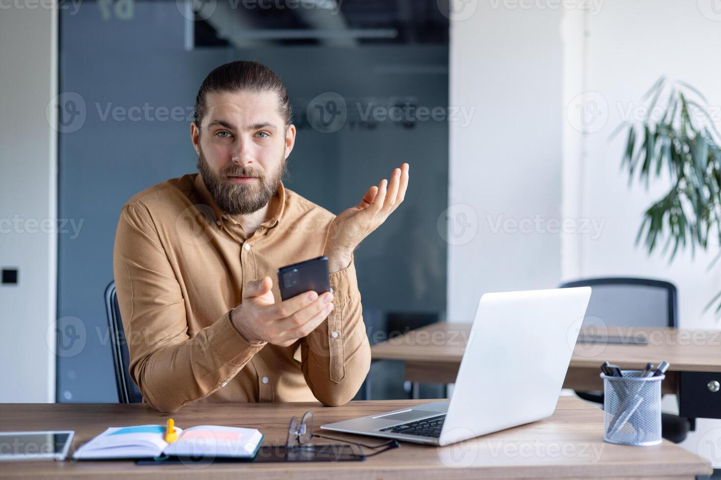 Professional office setting with a man holding a smartphone while working on a laptop. Modern workspace showing casual workwear and productivity tools like notebooks and writing utensils. photo