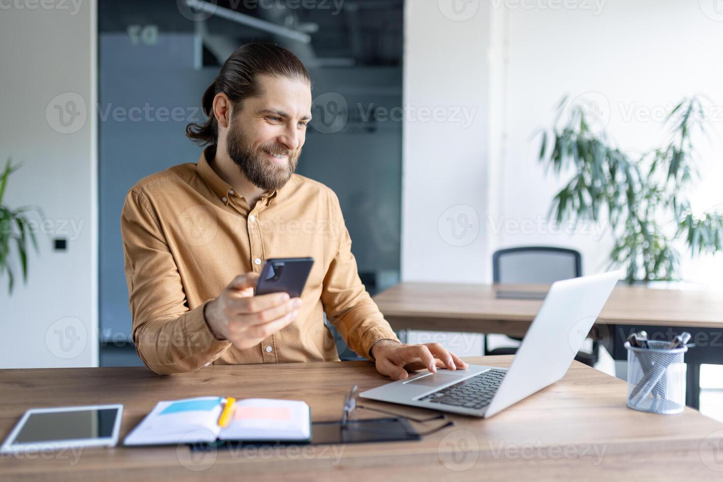 A modern office worker multitasking using a phone and laptop at a wooden table in an office. The scene conveys productivity and professional engagement with technology in a corporate environment. photo