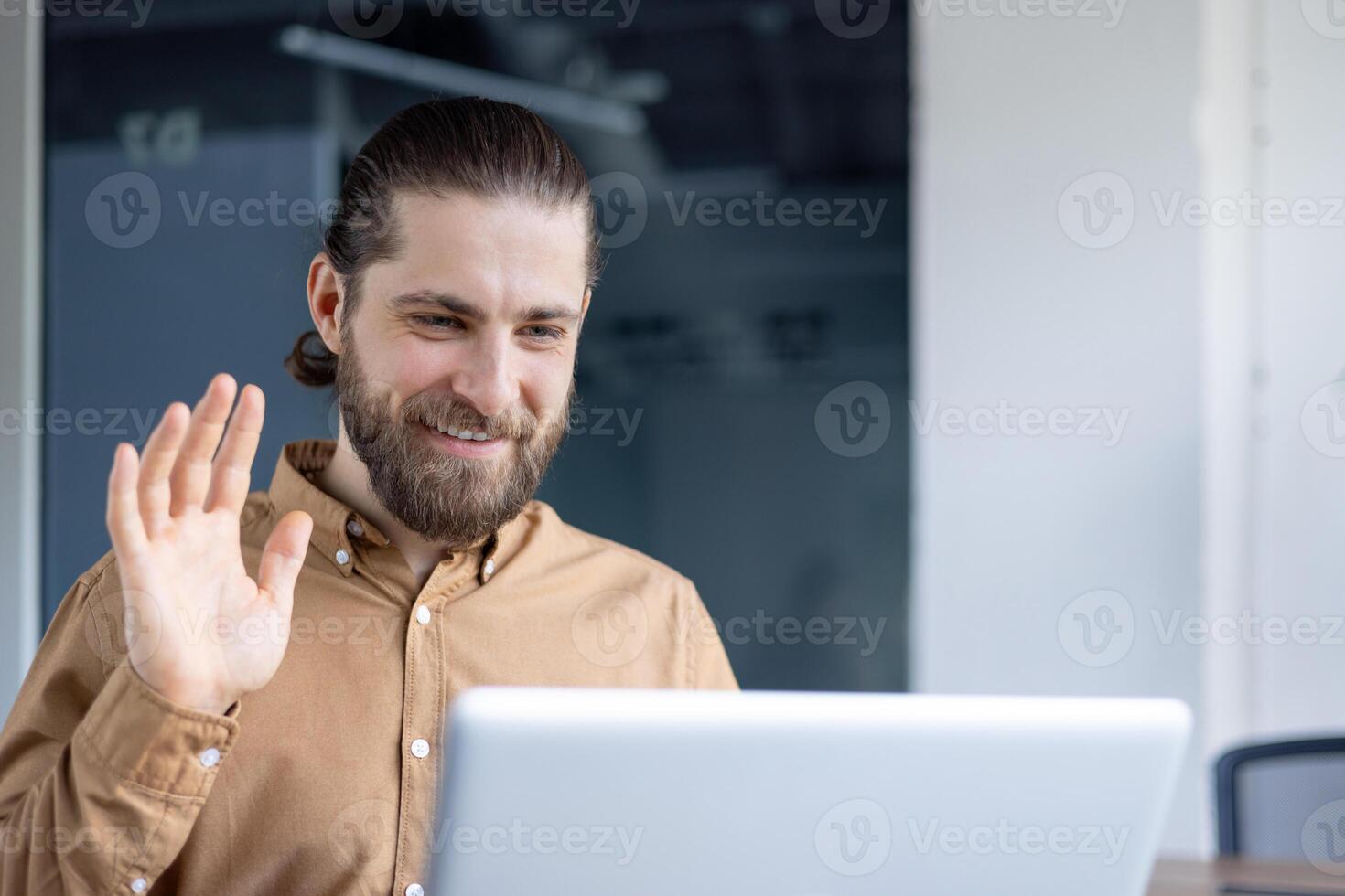 A bearded man in a casual shirt participates in a call, waving and smiling warmly at the screen. Ideal representation for communication, remote work, or online professional interactions. photo