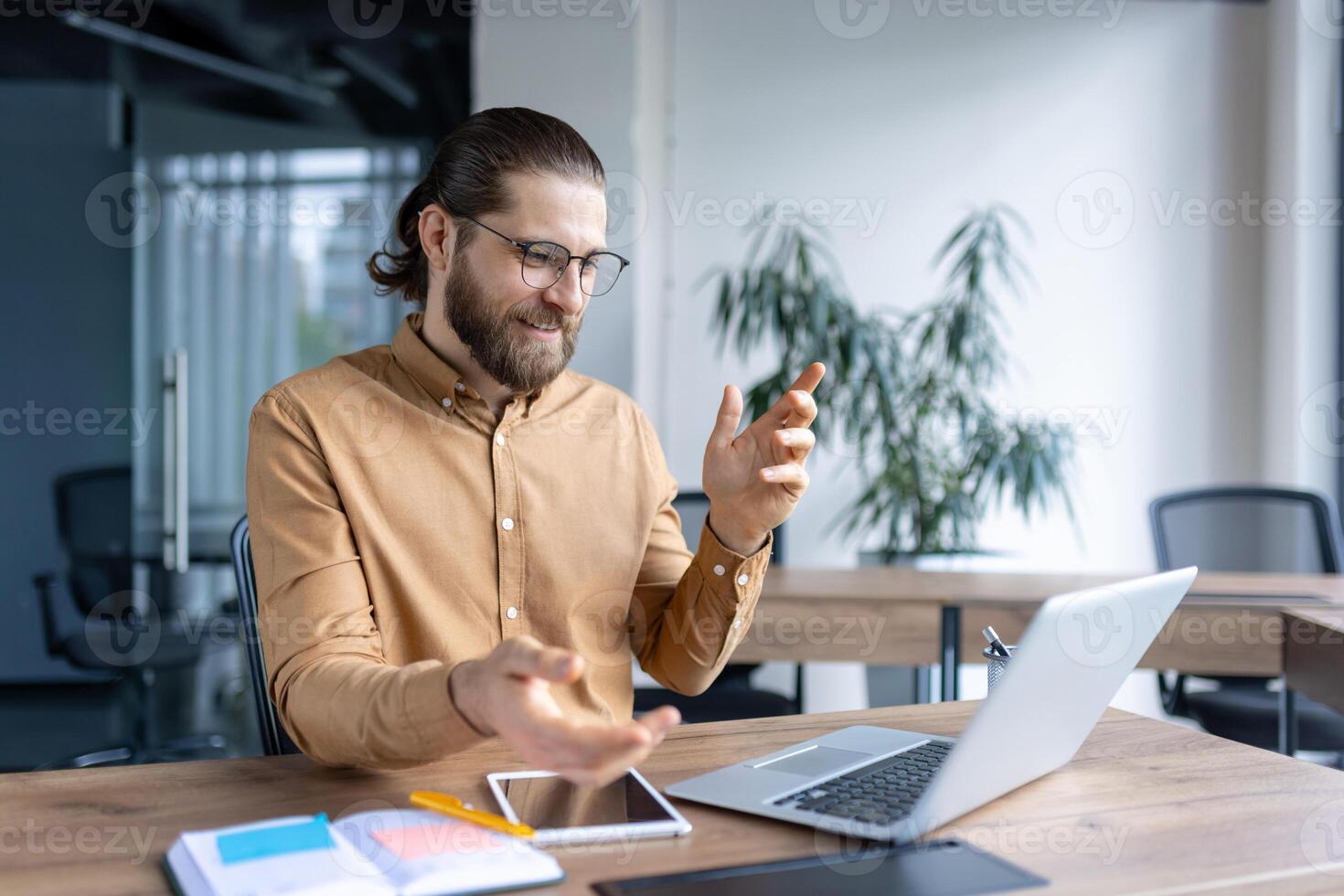 Young professional man communicating through a virtual meeting on his laptop at an office desk, with a smartphone and notebook. photo