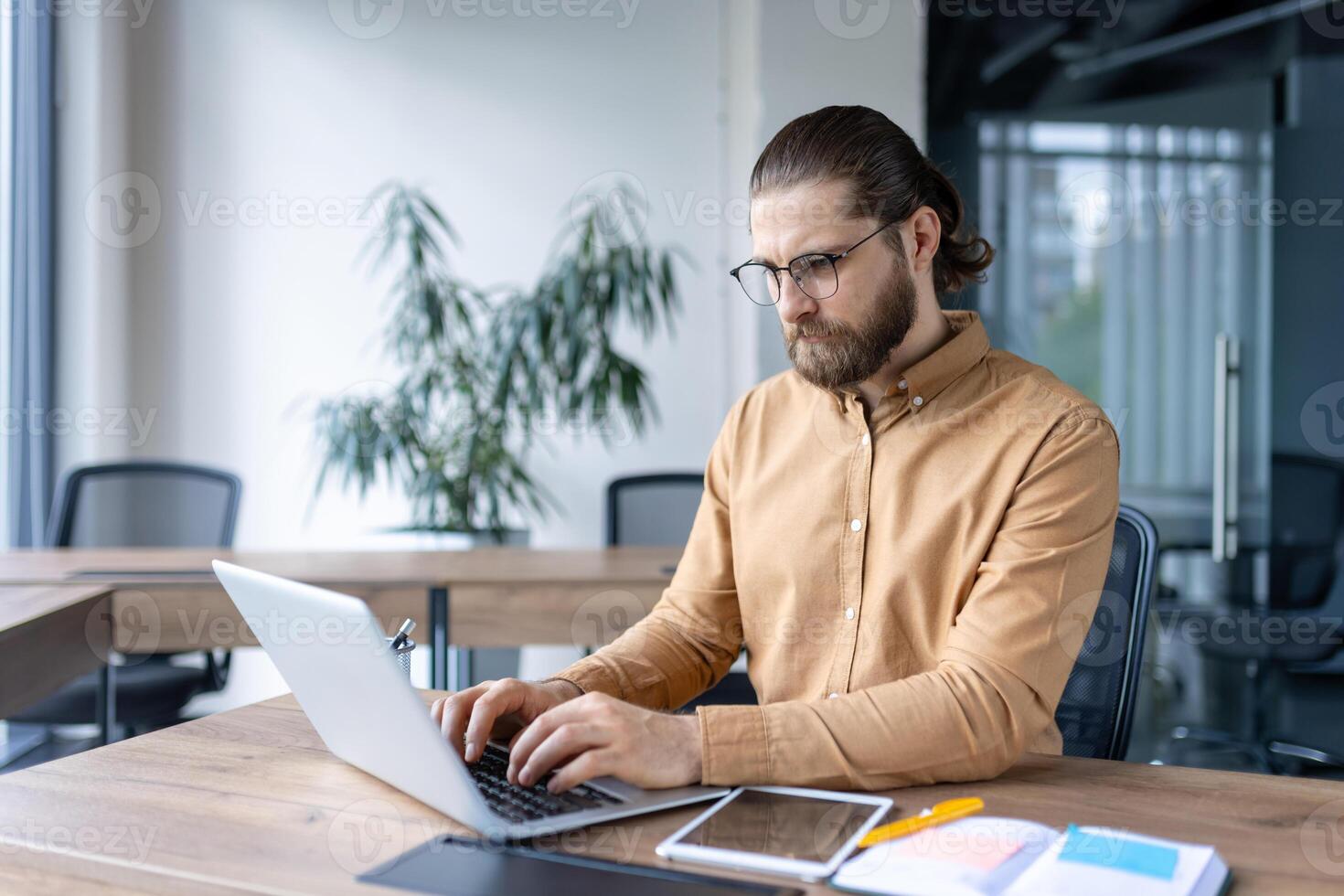 A professional male employee concentrates on his work using a laptop in a bright office setting, exuding productivity and focus. The workspace includes modern furniture and a calm ambiance photo