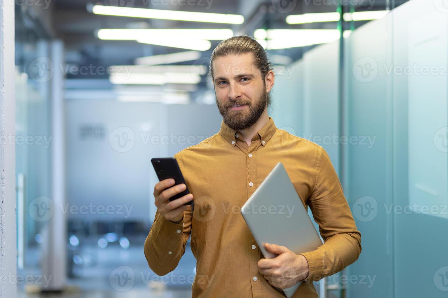 Young professional dressed in business casual attire holds a laptop and smartphone while standing in a modern office environment, showcasing confidence and tech-savviness. photo