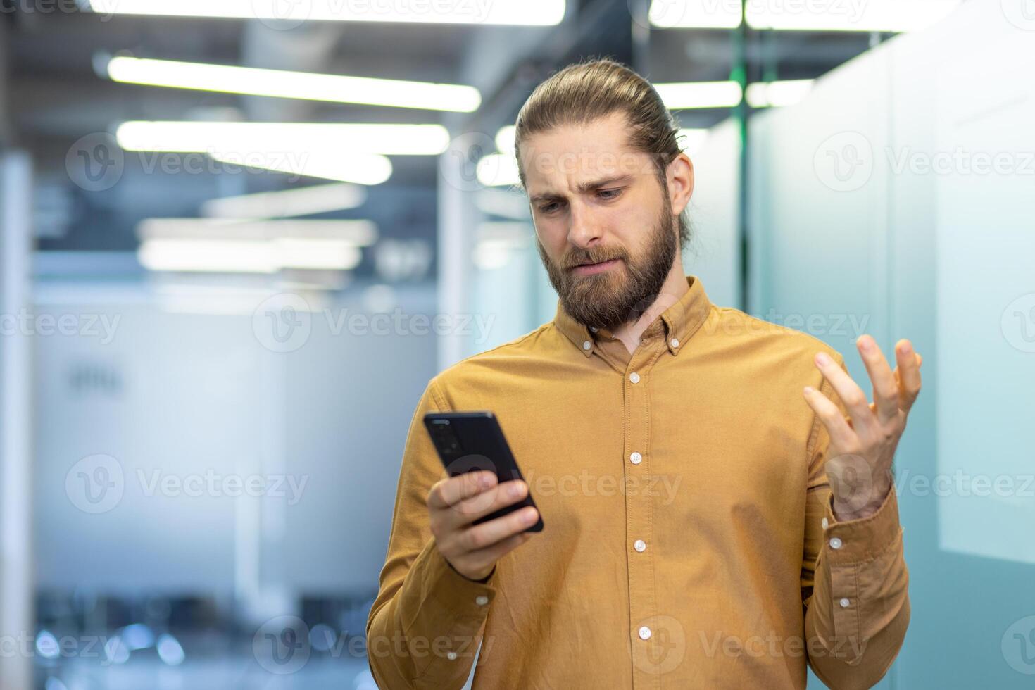 A man with a confused expression holds a smartphone, seeming frustrated or puzzled, in a bright, modern setting. photo
