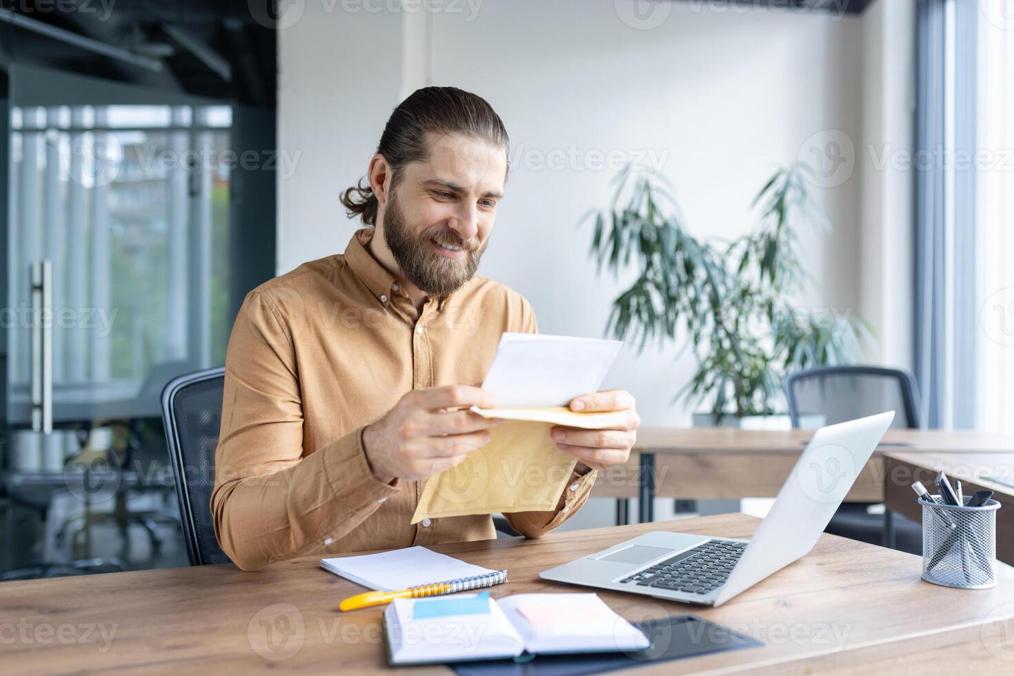 A businessman sits at his desk, smiling as he opens an envelope, amidst a professional environment. Perfect for concepts related to business, communication, office ambiance, and workplace joy. photo
