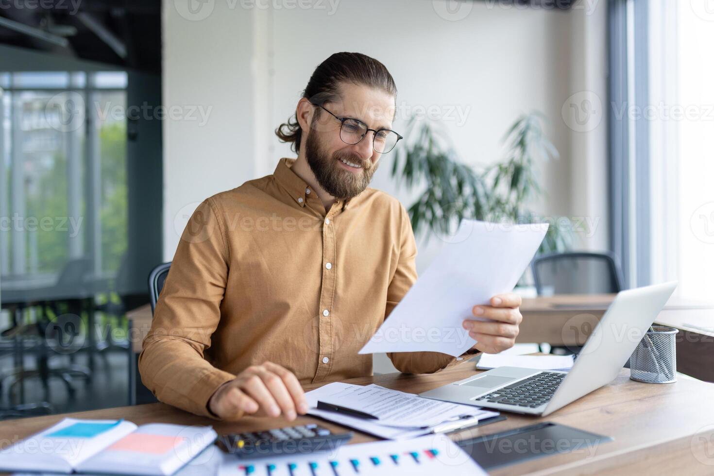 A focused professional reviews paperwork while working on financial or business tasks at a desk with a laptop and calculator, suggesting determination, productivity, and a modern office environment. photo