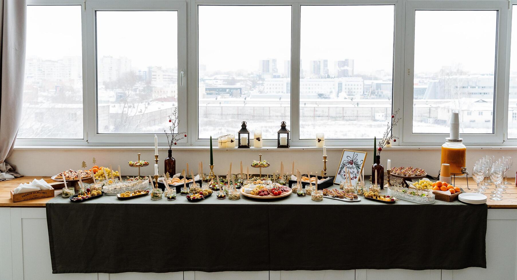 In an inviting home, a long, beautifully set table is filled to the brim with an array of delicious food and refreshing drinks, perfectly positioned in front of a large window photo