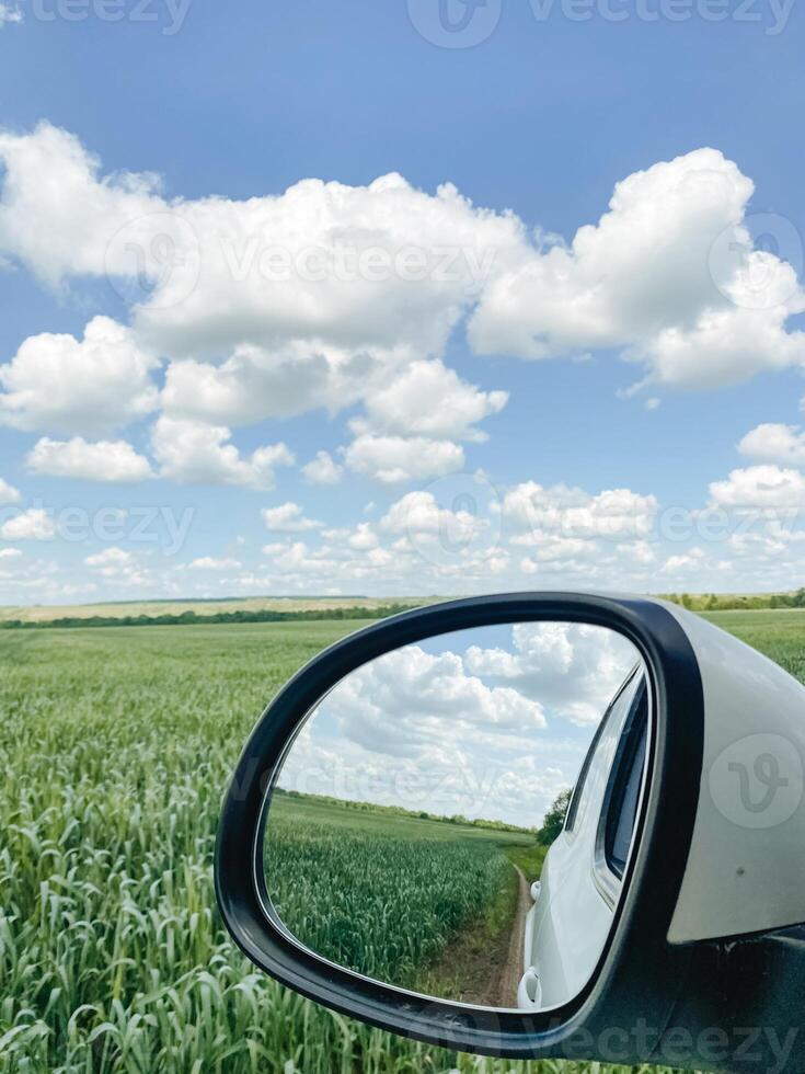 A cars rear view mirror effectively reflects a beautiful field along with some fluffy clouds present in the sky, capturing a serene and picturesque automotive experience photo