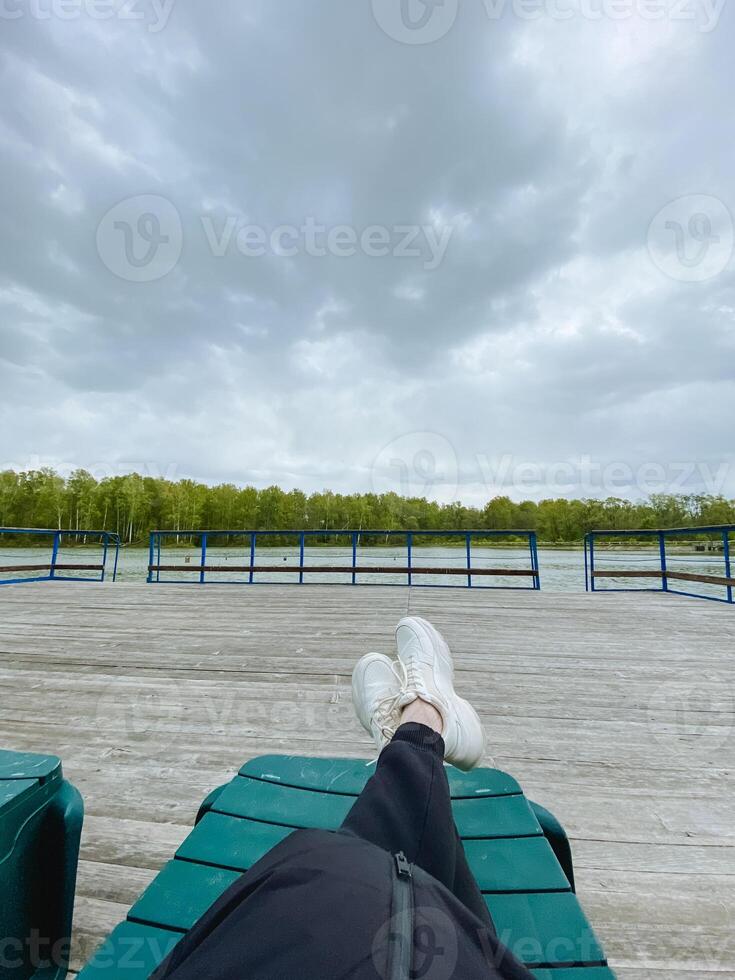 A person is comfortably lying back on a bench with their feet elevated, enjoying the view of the sky above, surrounded by fluffy clouds and the lush green grass all around them photo