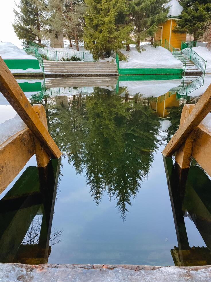 A beautifully constructed wooden bridge gracefully spans a serene body of water, while the trees nearby are perfectly mirrored in the calm surface of the water, creating a picturesque view photo