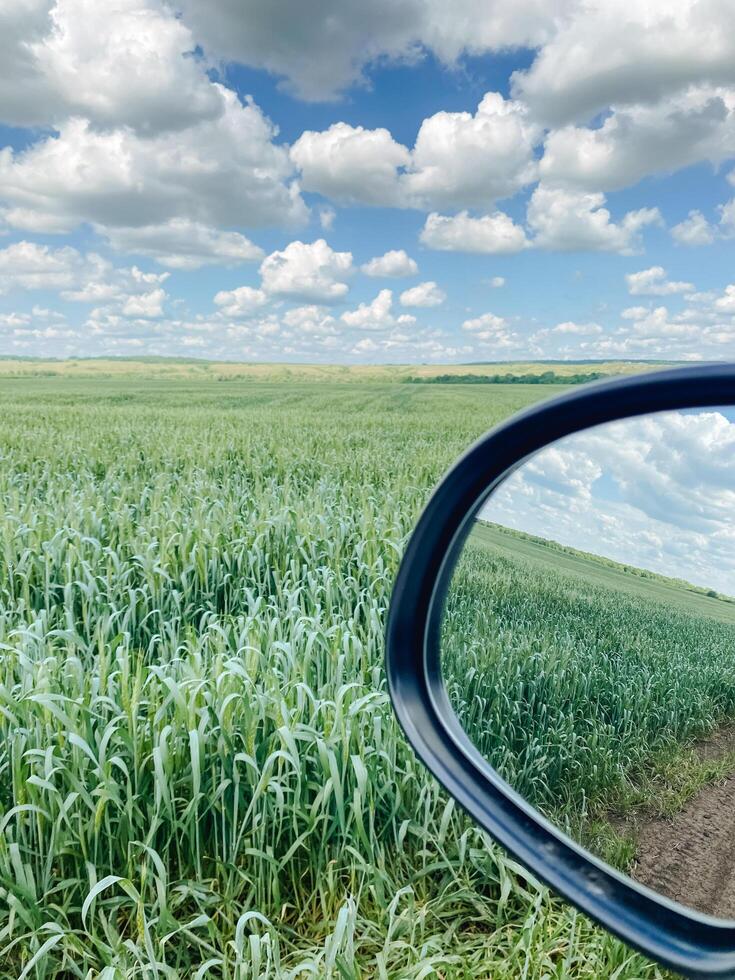 The rear view mirror captures the road behind me and beautifully reflects a stunning field filled with vibrant green grass and fluffy white clouds hovering in a calm, serene blue sky photo