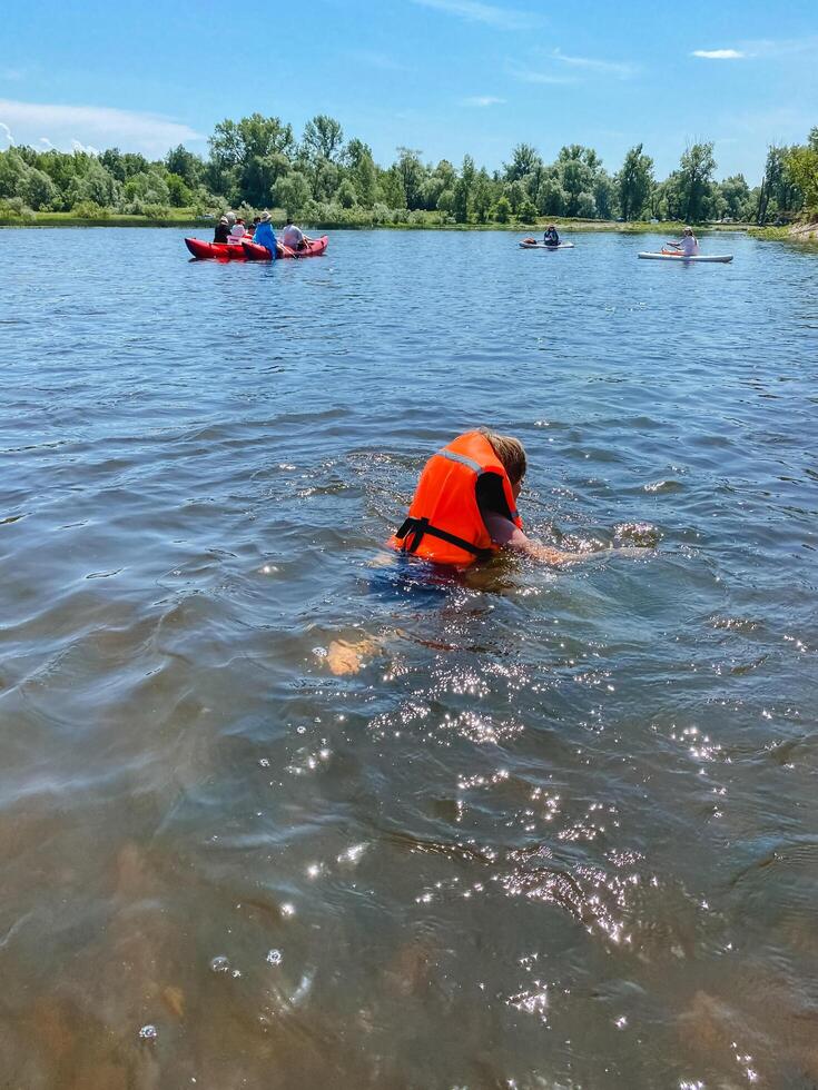 A cheerful person in a bright orange life jacket swims joyfully in a sparkling lake, enjoying the cool water and picturesque nature under a clear blue sky with a few fluffy clouds photo