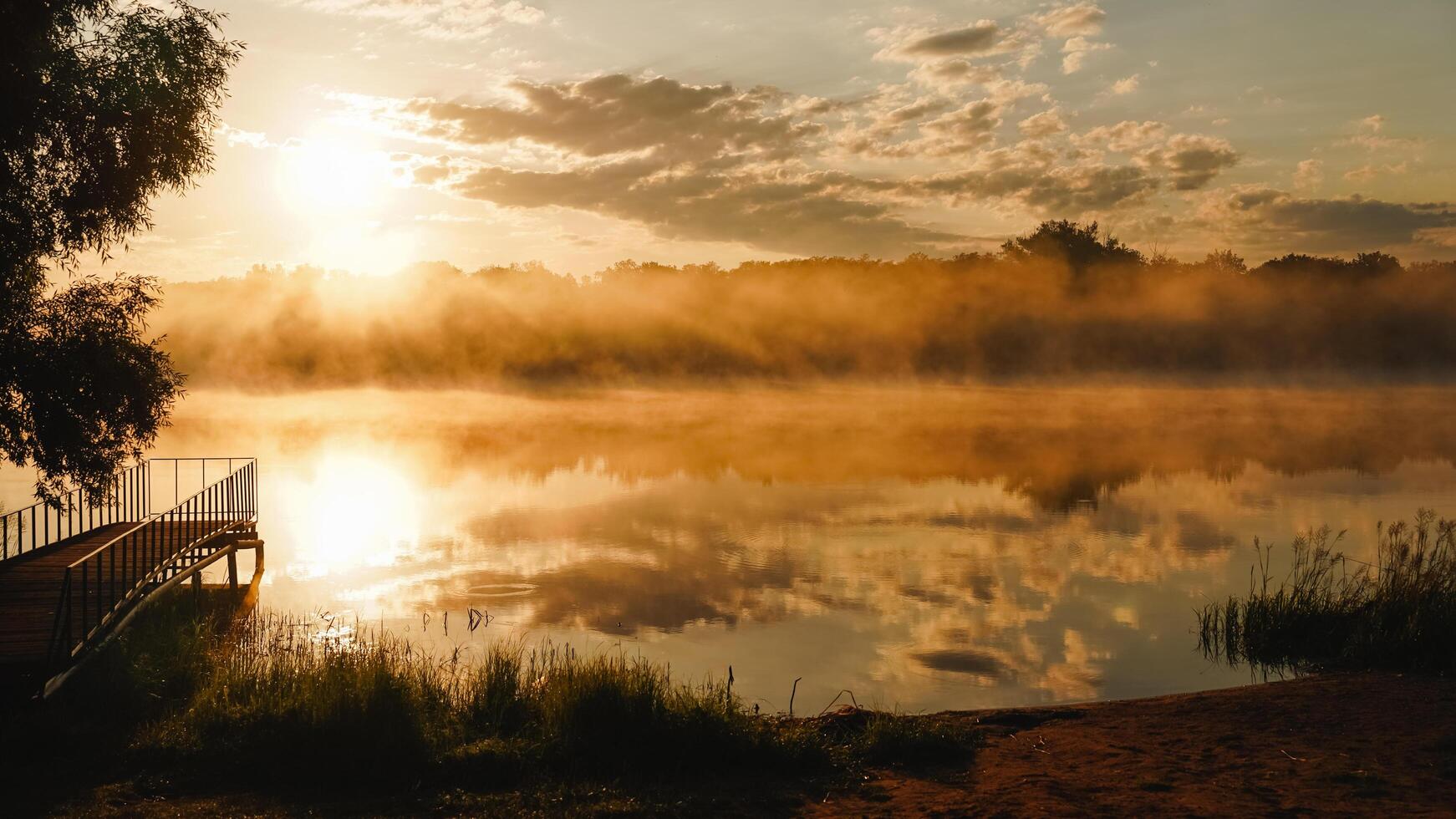 As the day comes to a close, the sun begins its descent, casting a warm glow over a misty lake where a wooden dock stands prominently in the foreground, creating a serene atmosphere photo
