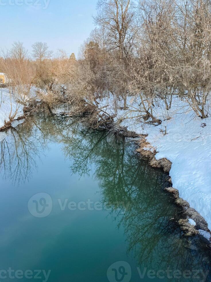 A beautiful river flows gracefully, adorned with snow on its tranquil shores, while trees stand tall in the background, creating a serene and picturesque natural landscape photo
