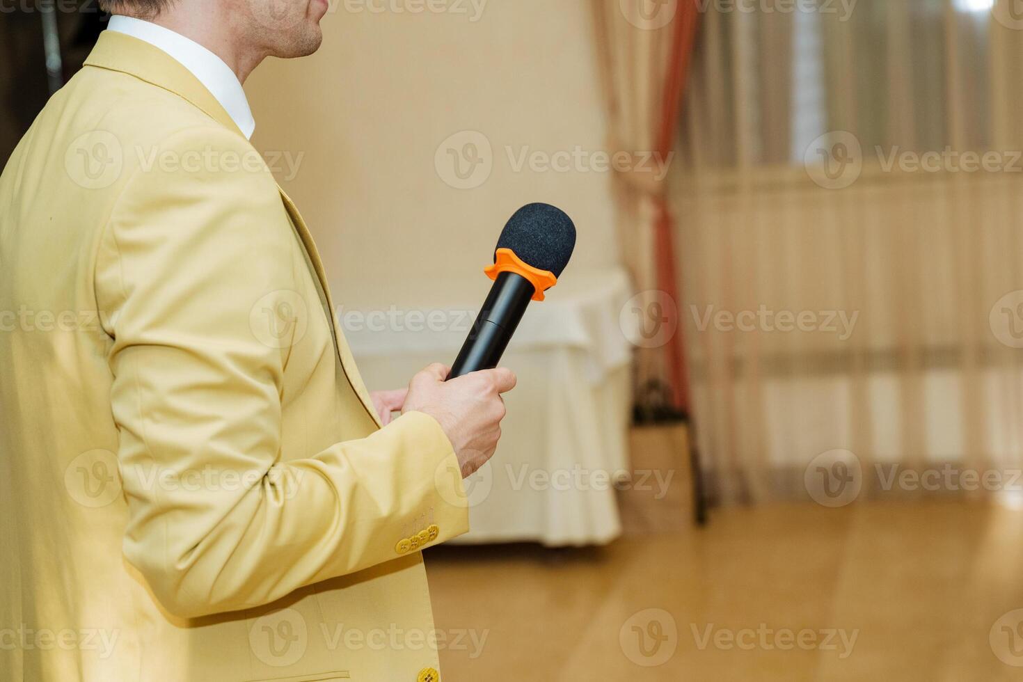 A man in a yellow jacket is standing and holding a microphone, probably giving a speech at a formal event. The setting includes a wooden flooring and a public address system photo