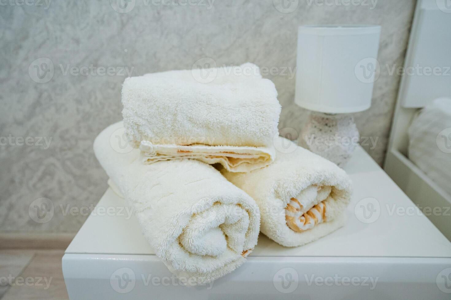 A sleek white table, situated next to a warm and cozy lamp, holds three neatly rolled towels, which enhances the clean and inviting atmosphere, promoting both comfort and stylish elegance photo