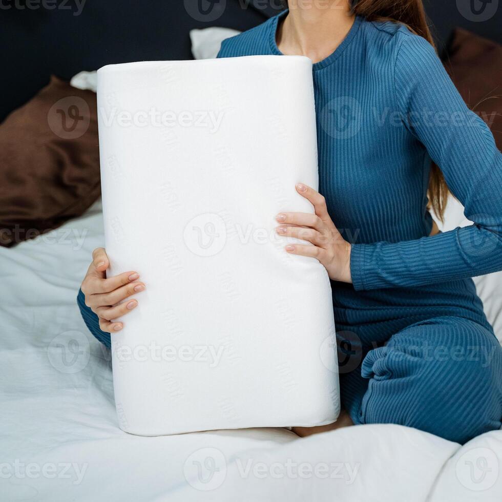 A casually dressed woman comfortably relaxes on her cozy and inviting bed, holding a soft, fluffy white pillow in her lap, which adds to the warm and welcoming atmosphere surrounding her photo