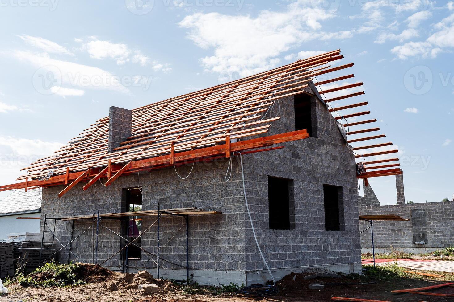A brick house is currently under construction, featuring a wooden roof that is being carefully placed. The project site is surrounded by various plants and an expansive land lot photo