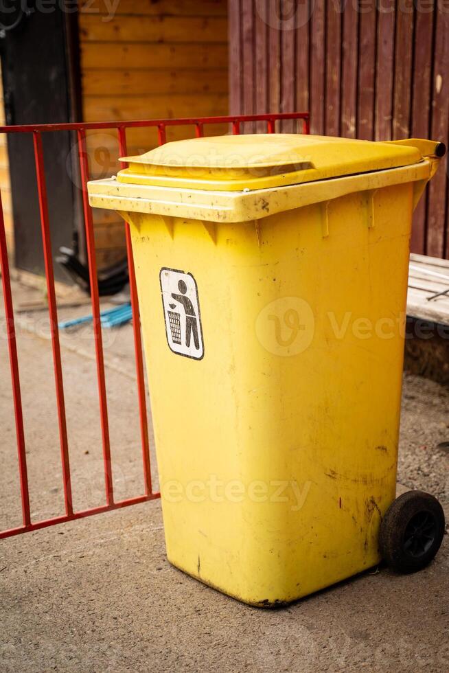 This is a bright yellow trash bin on wheels placed strategically in an urban setting for easy access and use photo