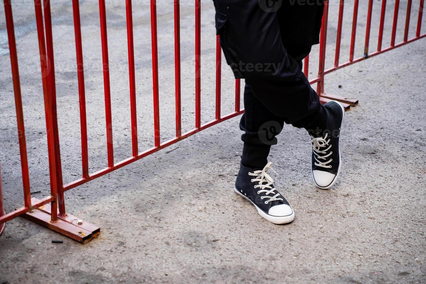 An individual is standing by a bright red fence, casually crossing their legs in an eyecatching pose that captures the attention of those nearby, enhancing and elevating the overall scene photo