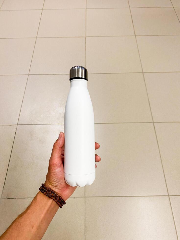 A person is standing, holding a white plastic water bottle with their fingers wrapped around it. It is clear the bottle contains liquid. The scene is likely indoors on a floor surface photo