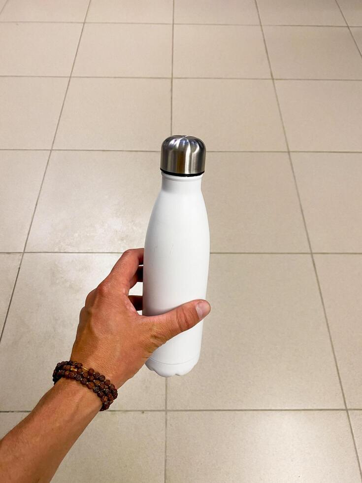 A person is standing on the flooring, holding a white plastic water bottle. It resembles a drinkware item. Keywords include fluid, gesture, gas, liquid, thumb, and human leg photo