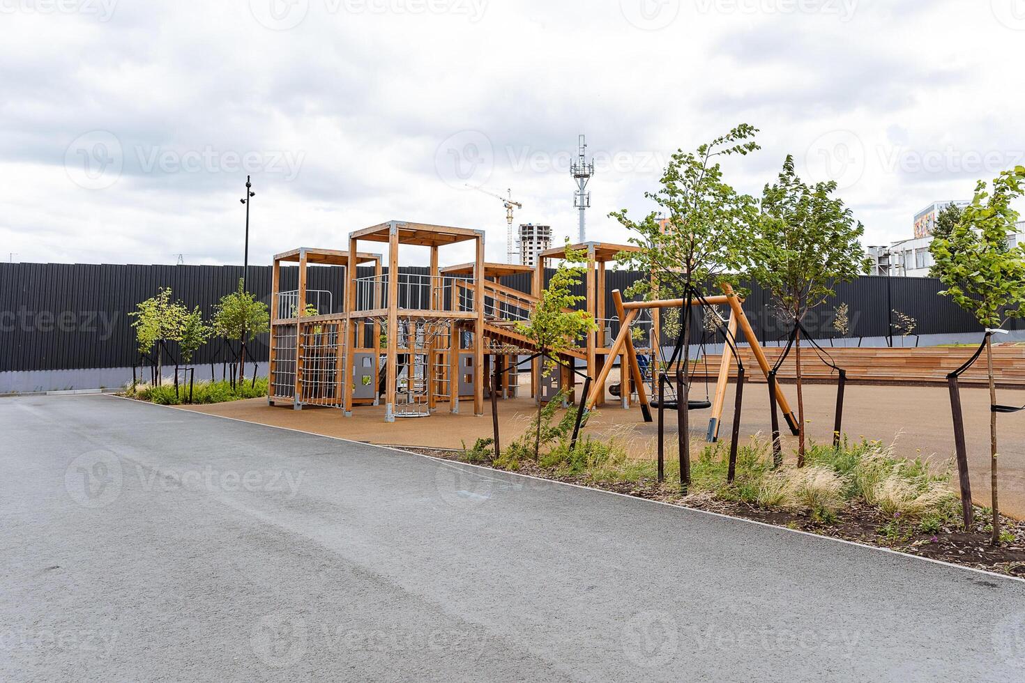A wooden playground is presently located on the side of a bustling road, surrounded by various elements of urban design and nature that create an interesting visual landscape photo
