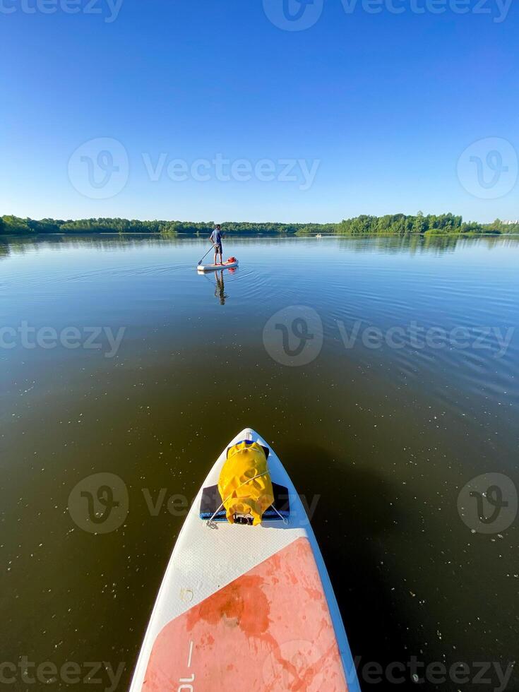A person leisurely paddles on a standup paddleboard in a serene lake surrounded by nature under a clear blue sky during the daytime, enjoying the peaceful natural environment photo