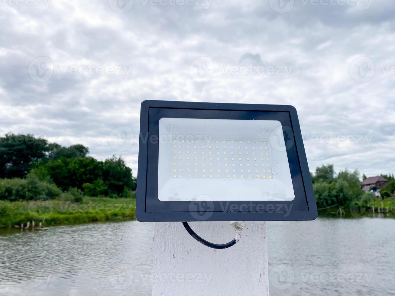 A powerful flood light is positioned on top of a tall white pole, which stands next to a serene body of water, likely a lake that reflects the surrounding landscape beautifully photo