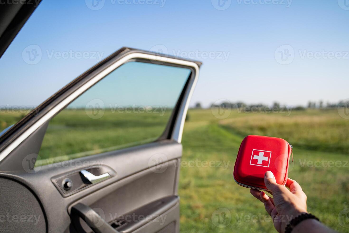 A hand is holding a first aid kit by the open car door set against a beautiful green field landscape photo