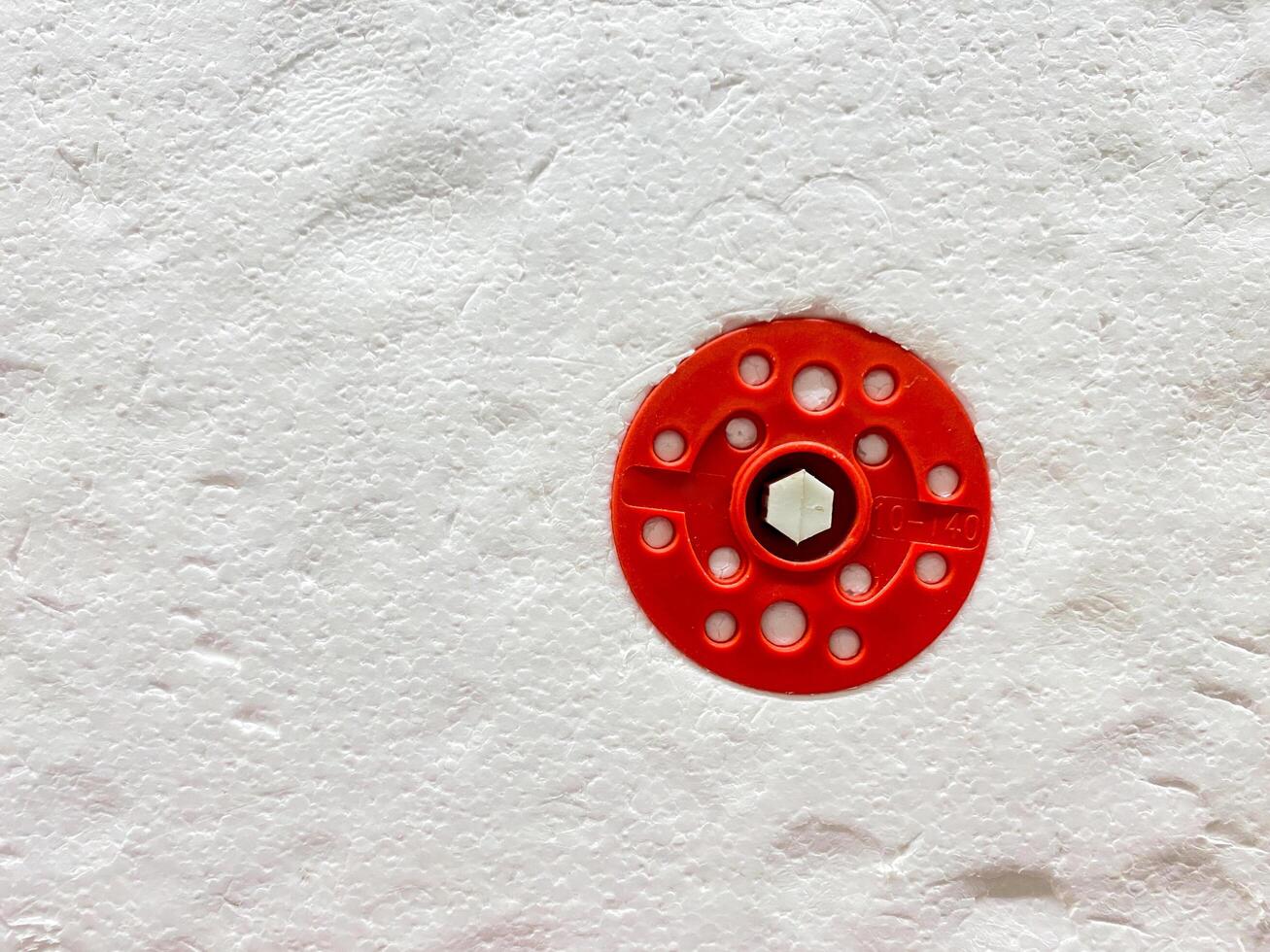 On a pristine white wall, there is a striking red circle that has multiple holes punctuated within its design, creating a visually interesting contrast with the background surface photo