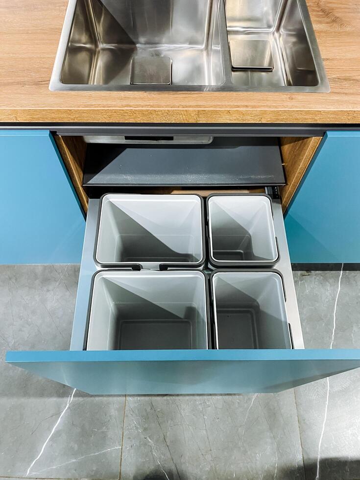 In a kitchen space, there is a sink prominently positioned, and right below it, you can find a drawer that is filled with trash bins that are designated for waste disposal photo