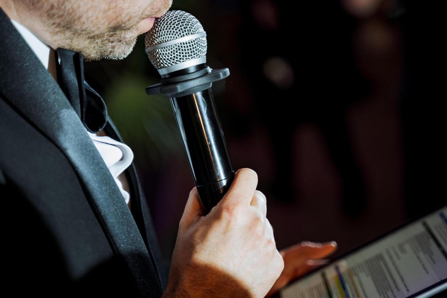 Man in formal wear gestures with microphone while reading paper photo