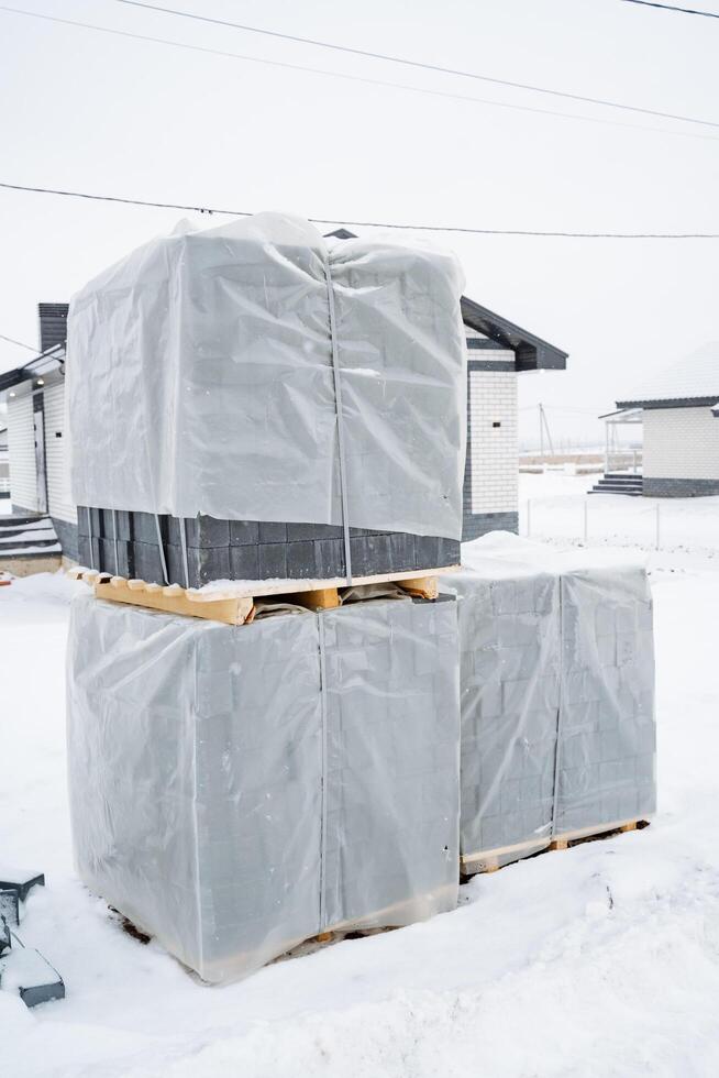 A stack of neatly wrapped bricks rests on a wooden pallet, surrounded by a thick layer of untouched snow, creating a stunning contrast against the serene winter landscape photo