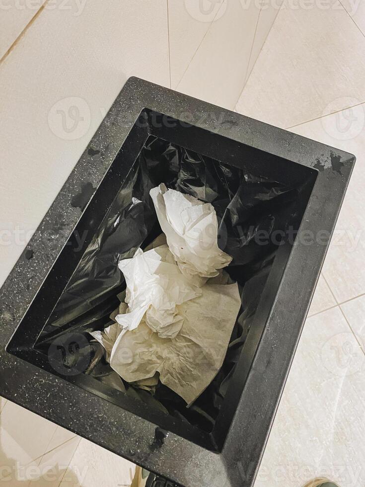 An overflowing black trash can filled with used tissues sits awkwardly on the tiled floor, creating a messy look that contrasts sharply with the otherwise clean surroundings photo
