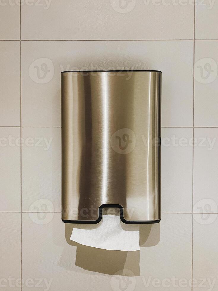 A shiny stainless steel paper towel dispenser, designed in a sleek rectangular shape, is hanging on a tiled wall that adds a touch of modernity to the restroom or kitchen area photo