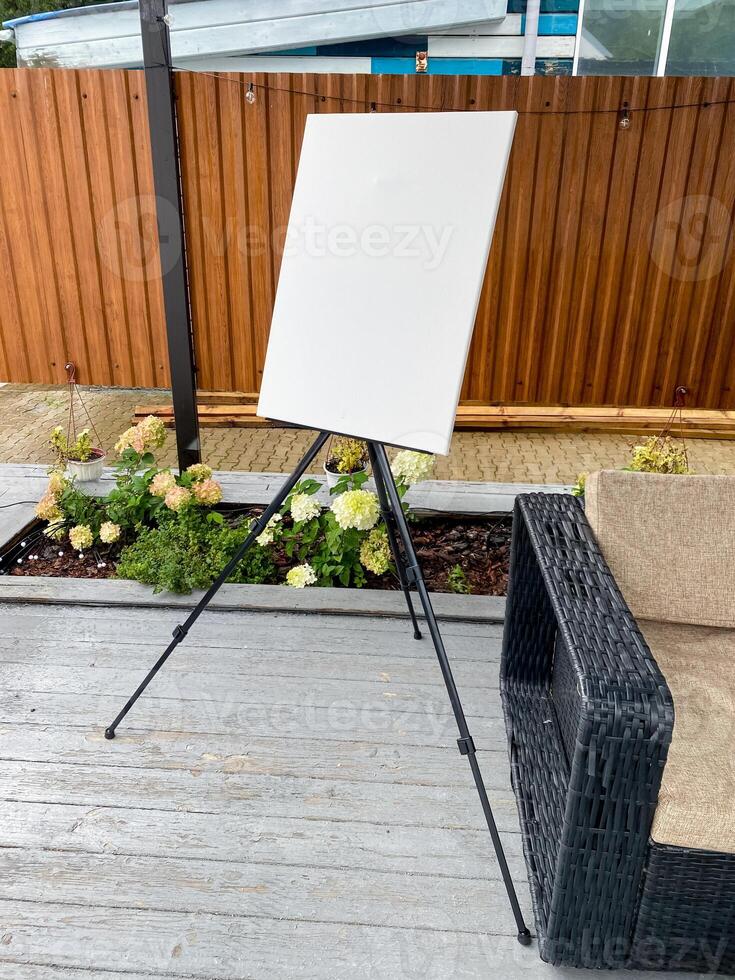 There is an easel holding a white canvas set on a deck beside a chair photo