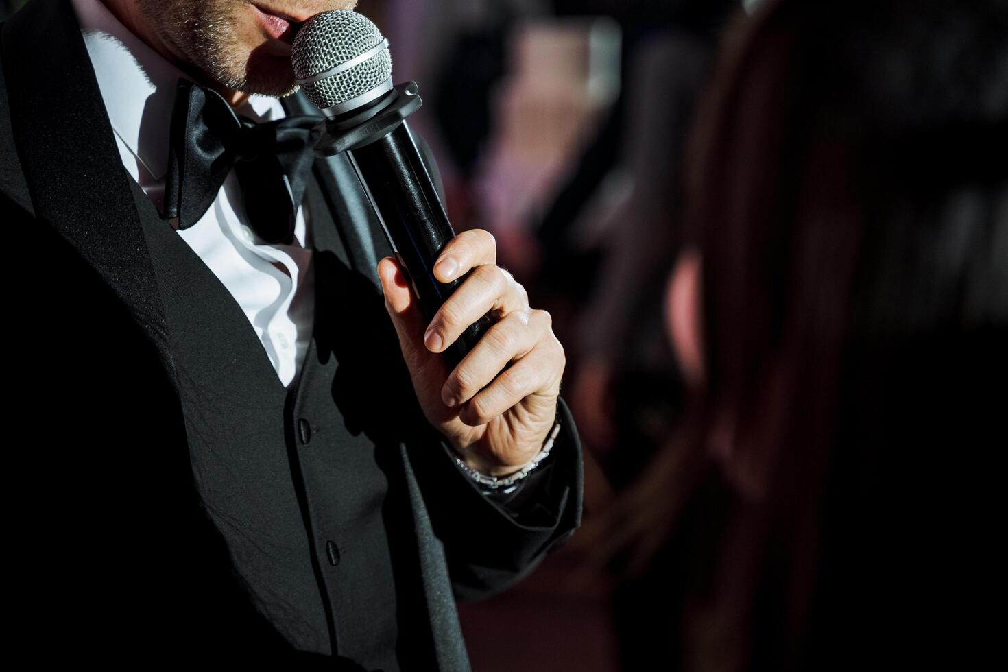 A man in formal wear gestures with microphone towards a woman at an event photo