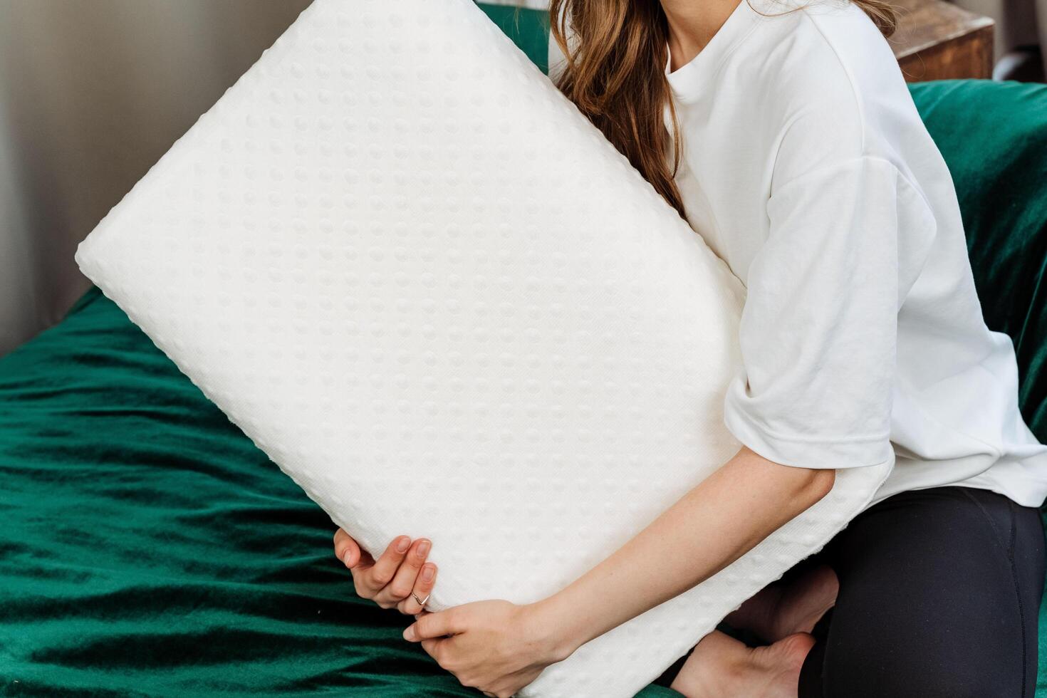 A young woman is comfortably seated on a green bedspread, holding a white memory foam pillow. The setting exudes a sense of calm and relaxation in a cozy indoor environment photo