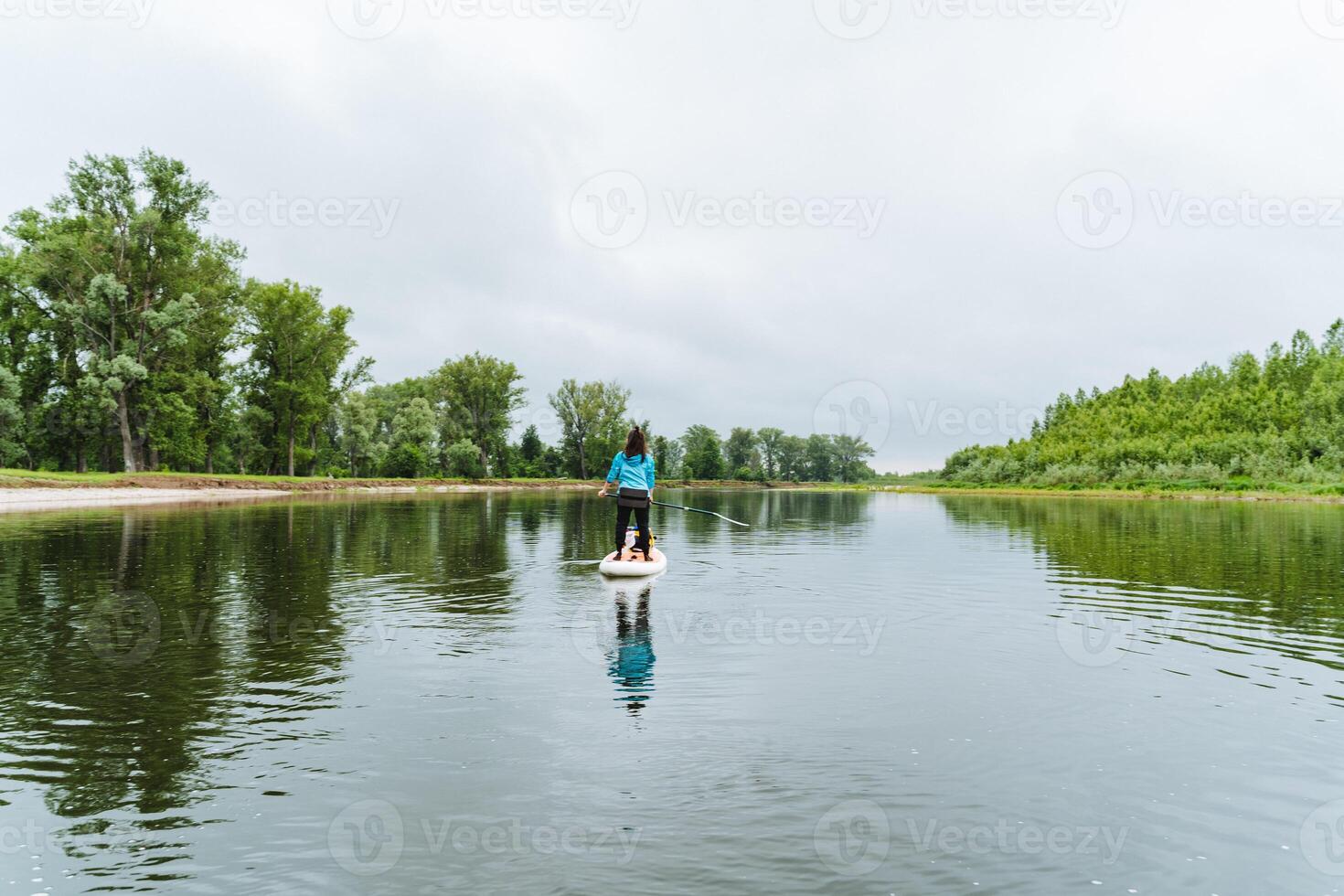 A woman calmly paddle boards down a serene river, amidst beautiful nature, under clear skies. The scene is peaceful and tranquil, surrounded by lush greenery and natural beauty photo