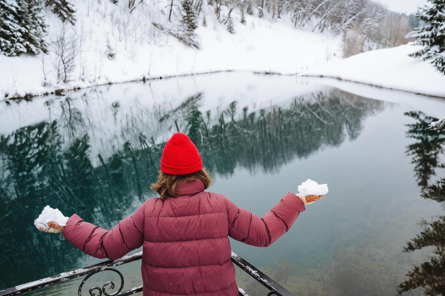 A woman, elegantly dressed in a striking red hat and matching jacket, is standing in front of a beautiful, snowy lake that glistens under the winter sun, creating a picturesque scene photo