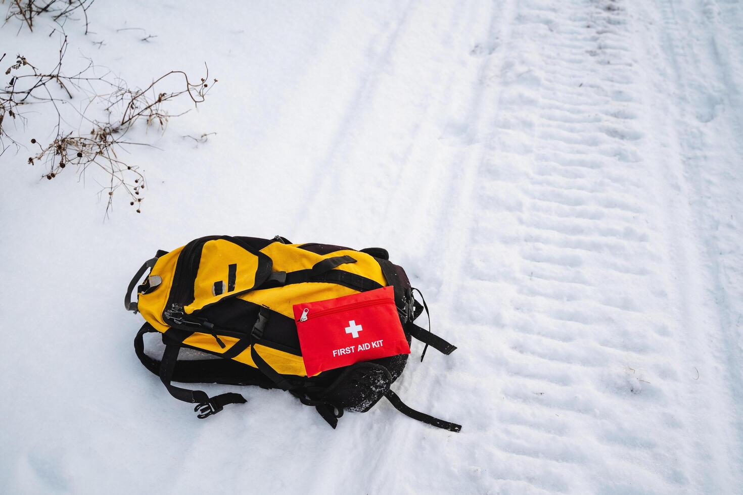 A bright yellow and black backpack is resting in the fluffy white snow, situated right beside a vibrant red first aid kit, creating a stark contrast in this winter landscape photo