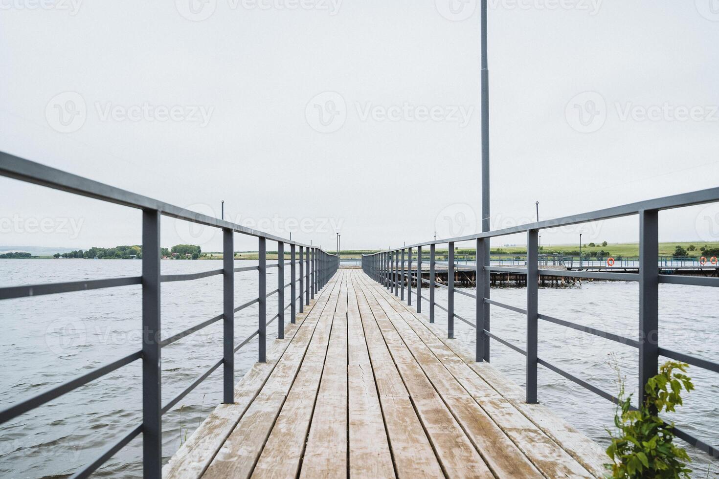 A wooden pier with a metal railing extends over calm water beneath a clear sky, forming a peaceful scene by the lake. It provides a serene spot for relaxation and contemplation photo